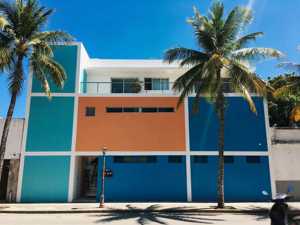 La fachada de un edificio en bloques de turquesa, naranja y azul, enmarcada por palmas bajo un cielo despejado.