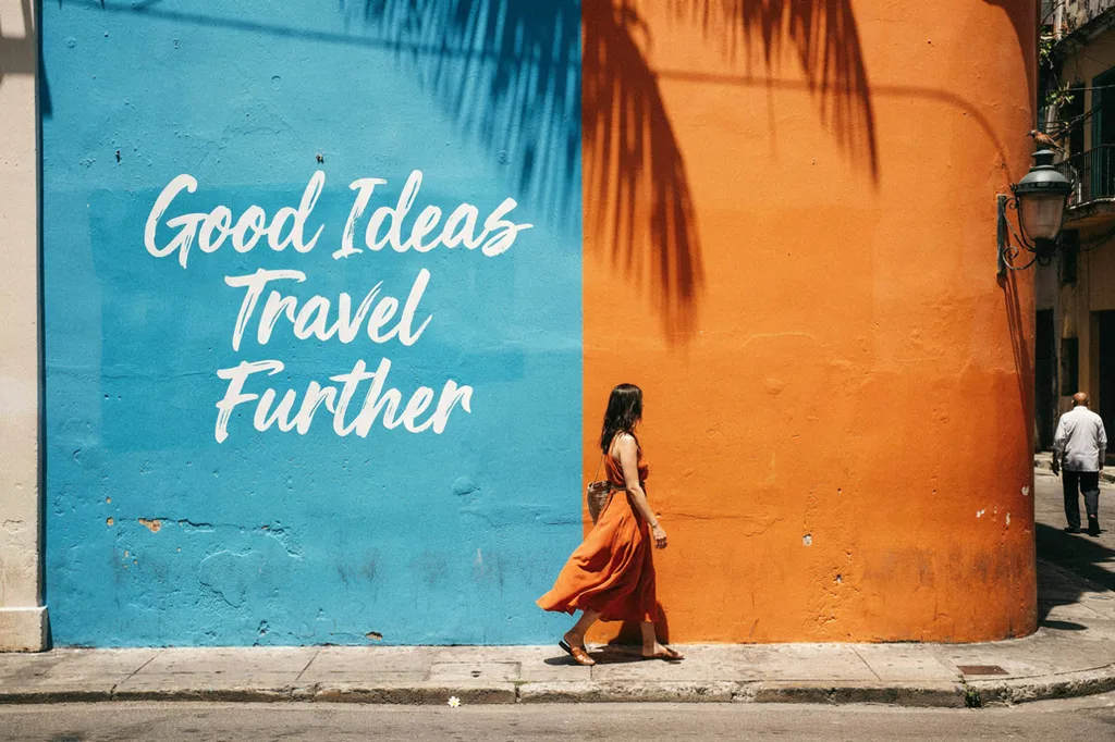 Un mural pintado a mano que dice “Good Ideas Travel Further” en una pared entre palmas.